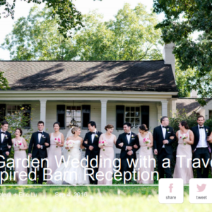 A wedding party stands in front of a white house on a sunny day. The group is dressed formally, with bridesmaids in light dresses and groomsmen in dark suits, all holding bouquets and smiling.