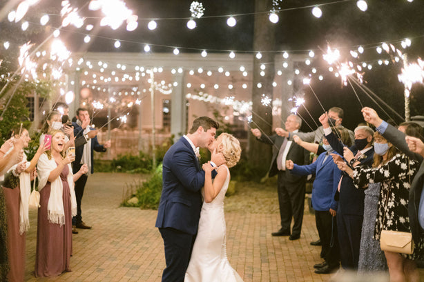 A bride and groom kiss under string lights as guests surround them holding sparklers. The scene is festive, with guests dressed formally and some wearing masks, creating a romantic celebration atmosphere.