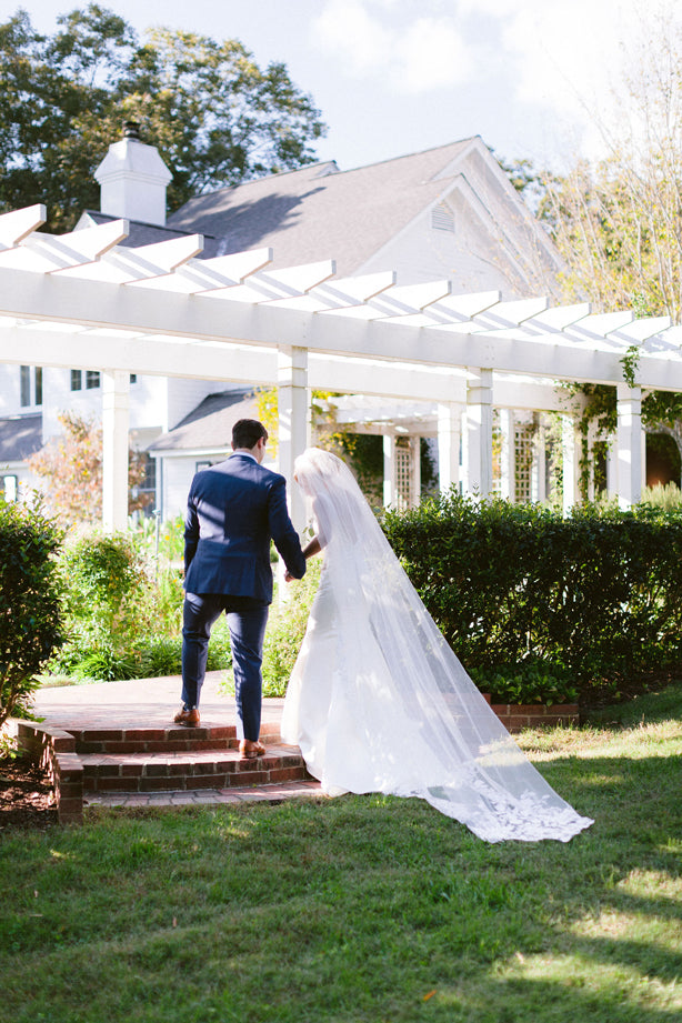 A bride in a long white gown and veil walks beside a groom in a blue suit, holding hands as they ascend brick steps toward a white pergola and a large house surrounded by greenery.