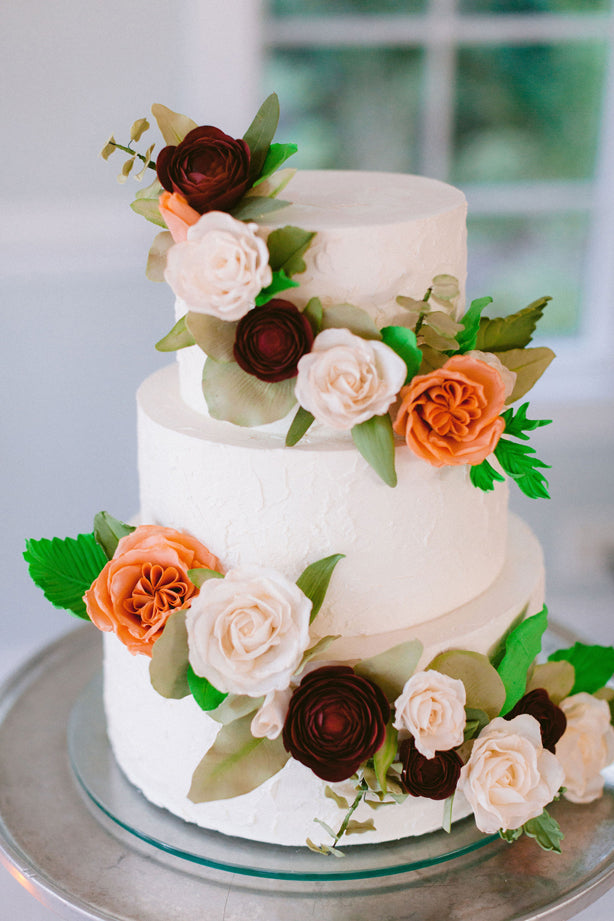 A three-tier white cake decorated with realistic orange, white, and burgundy flowers and green leaves, displayed on a silver cake stand in front of a window.