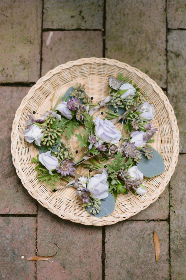 A round wicker tray holds several small floral boutonnieres with white roses, green leaves, and purple accents, placed on a red brick surface.