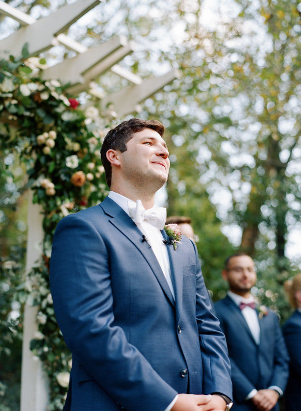 A groom in a blue suit and white bow tie stands and smiles outdoors under a floral arch, with groomsmen in matching suits blurred in the background. Trees and greenery are visible around them.