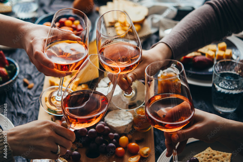 Four people clink glasses of rosé wine above a wooden board with cheese, grapes, and snacks, suggesting a celebratory meal or gathering.