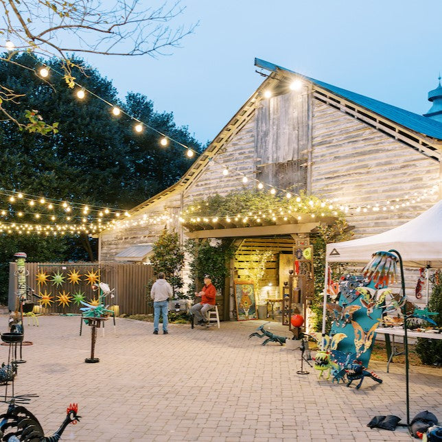 The Fearrington Folk Art Show, hosted by FEARRINGTON VILLAGE in a rustic barn adorned with string lights, will feature vibrant outsider art. Tall trees encircle the event, creating an enchanting atmosphere as visitors explore colorful sculptures during the Collector&#39;s Preview on February 21, 2025.