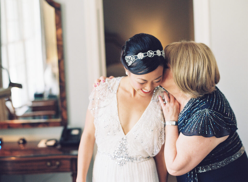 A woman in a beaded white dress smiles while an older woman in a dark, beaded dress leans in to whisper in her ear. They appear happy, sharing an intimate moment in a softly lit room.