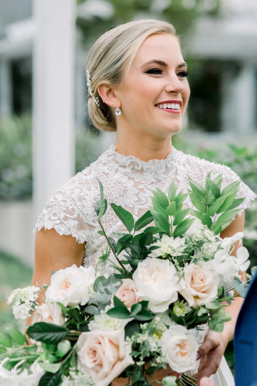 A smiling bride with blonde hair in an updo, wearing a lace wedding dress and pearl earrings, holds a bouquet of white and blush roses with greenery. The background is softly blurred with greenery.