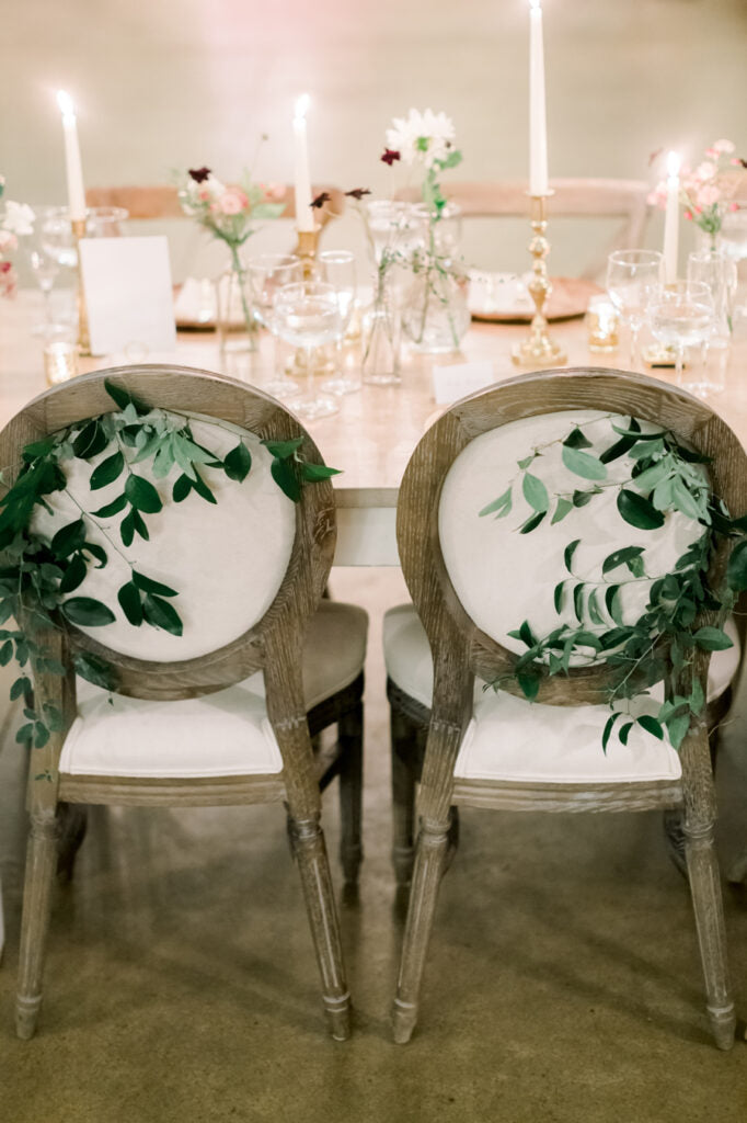 Two elegant wooden chairs with white cushions are decorated with green leafy garlands on their backs. The chairs are placed at a table set with flowers, candles, and glassware in a softly lit setting.