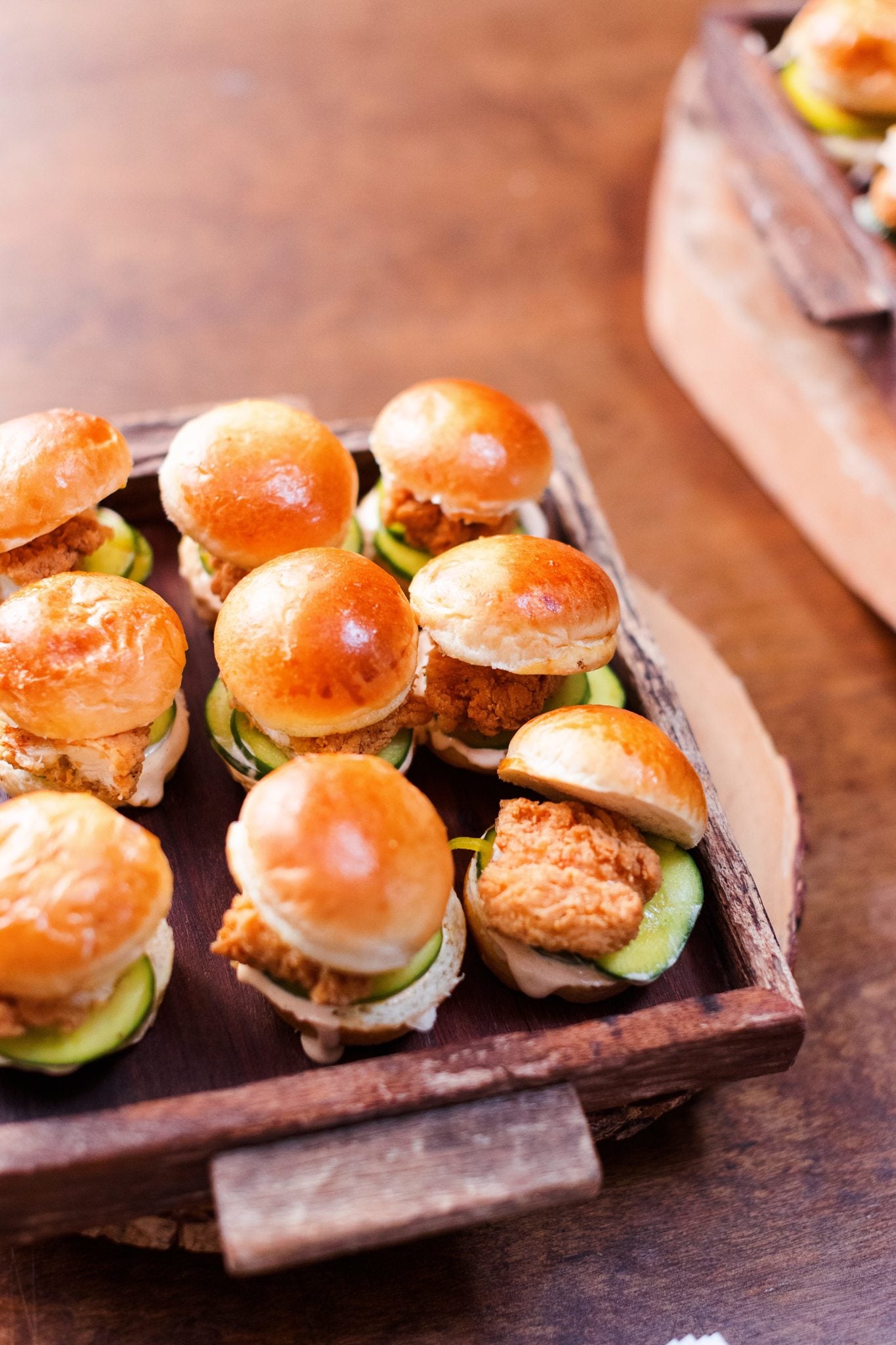 A wooden tray filled with mini sandwiches, each with a golden bun, a piece of fried chicken, cucumber slices, and sauce, arranged neatly on a wooden table.
