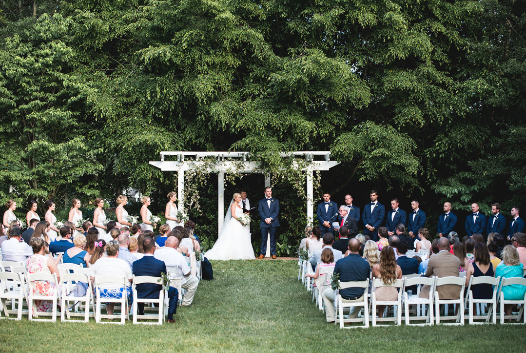 A bride and groom stand under a white arch during an outdoor wedding ceremony, surrounded by their wedding party and guests seated on white chairs, with lush green trees in the background.