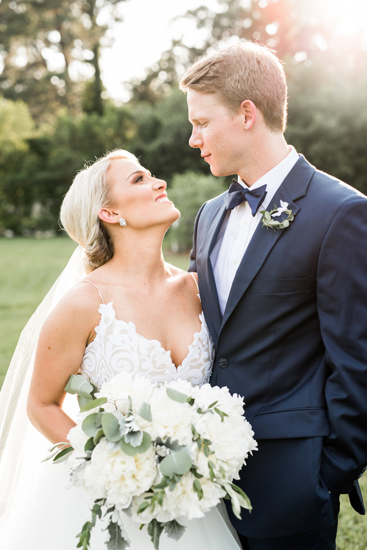 A bride and groom stand close outdoors, gazing into each other's eyes and smiling. The bride holds a white bouquet and wears a white dress and veil; the groom wears a navy suit and bow tie. Sunlight glows behind them.