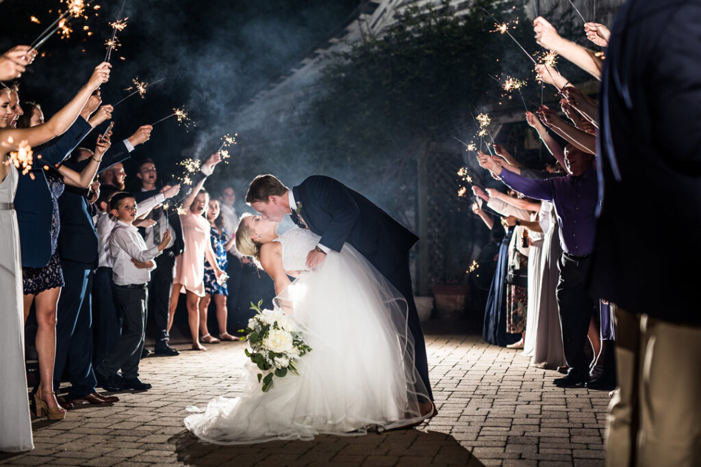 A bride and groom kiss while the groom dips the bride in the center of a walkway, surrounded by guests holding sparklers above them, celebrating at night. The bride holds a bouquet of white flowers.