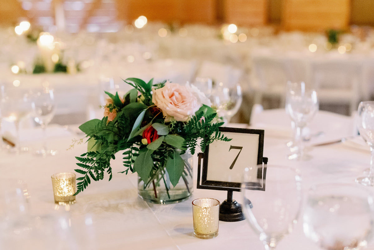 A table set for an event features a floral centerpiece with pink and red flowers and greenery, a table number 7 sign, empty wine glasses, and small gold votive candles on a white tablecloth.