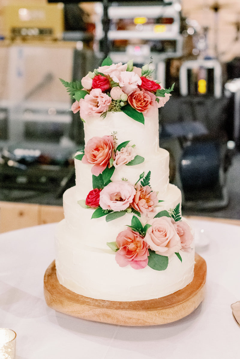 A three-tiered white cake decorated with pink and red flowers and green leaves, displayed on a wooden cake stand on a white tablecloth.