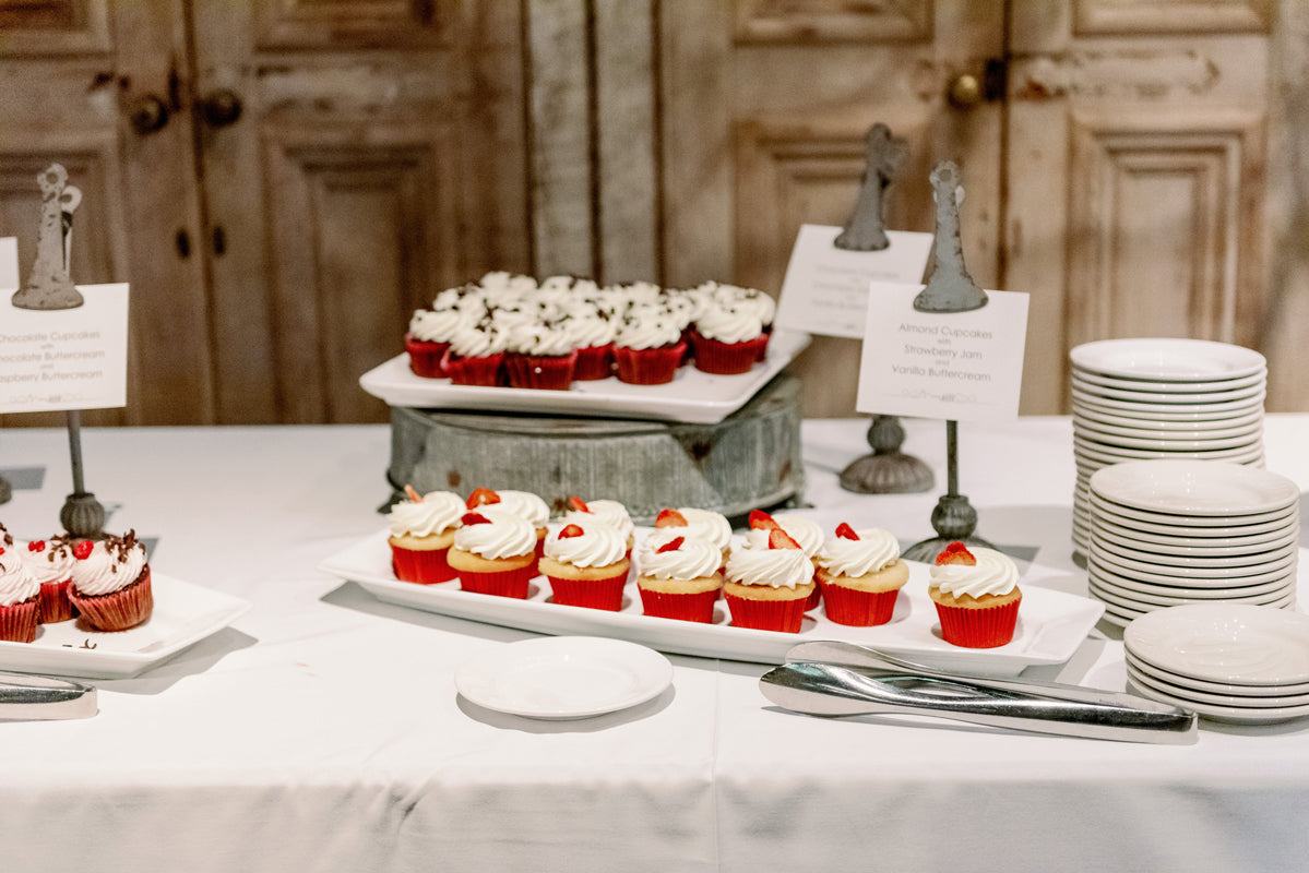A dessert table with red velvet cupcakes topped with white frosting and strawberries, arranged on trays next to stacks of white plates and utensils, with a rustic wooden background.