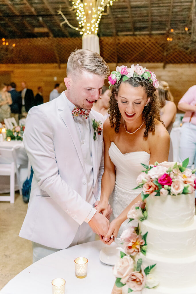 A bride and groom, both smiling, cut a white tiered wedding cake decorated with flowers. The bride wears a flower crown and strapless dress; the groom wears a light suit with a bow tie. The setting is warmly lit and festive.