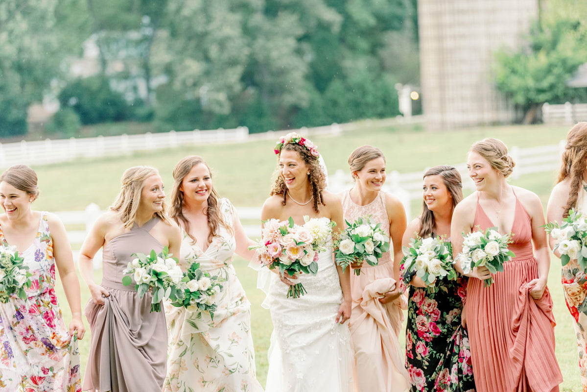 A bride in a white dress and flower crown walks outdoors with her bridesmaids, all holding bouquets and wearing colorful, floral or pastel dresses. They are smiling and laughing together on a grassy field.