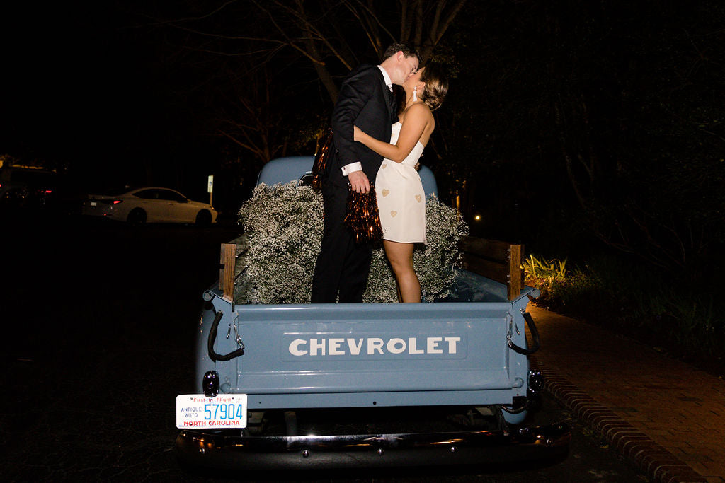 A couple dressed in formal attire stands kissing in the back of a blue vintage Chevrolet truck filled with flowers at night. The truck is parked on a street, and the scene is illuminated by soft lighting.