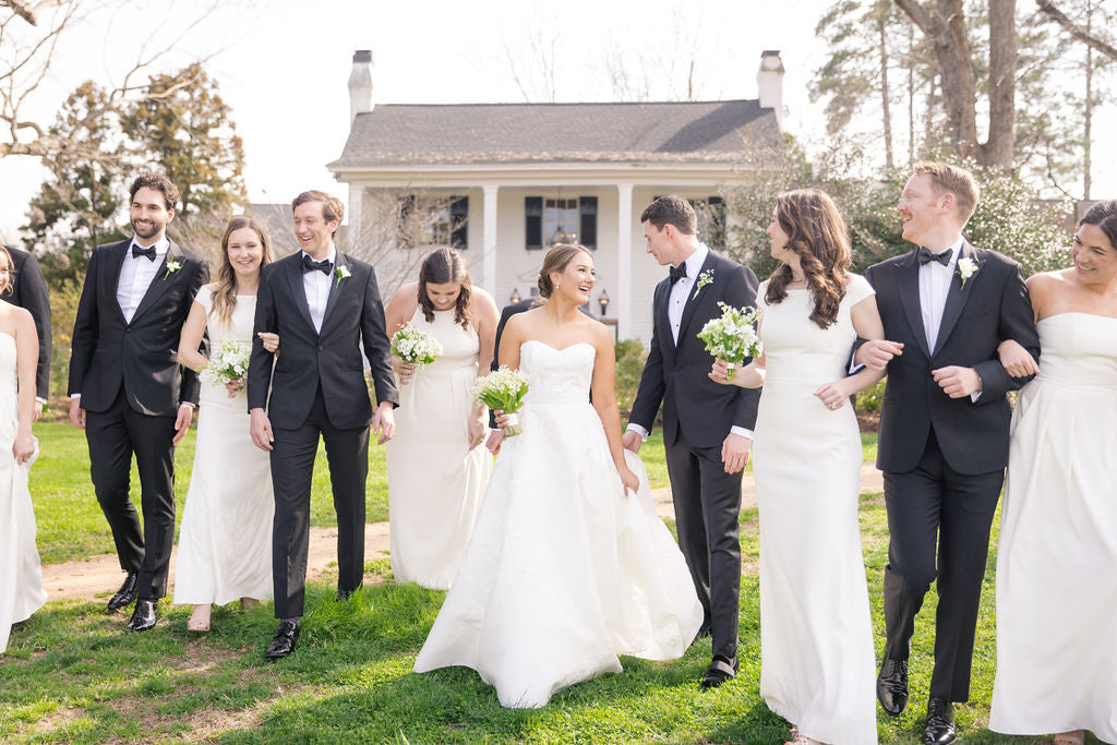 A joyful wedding party, dressed in black tuxedos and white dresses, walks together on a green lawn in front of a white house with columns on a sunny day. Everyone is smiling and holding small bouquets.