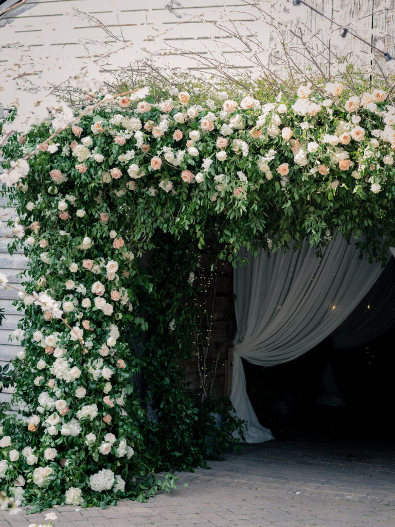 A large floral archway made of lush greenery and white and blush roses frames the entrance to a venue draped with white fabric, creating an elegant and inviting atmosphere.