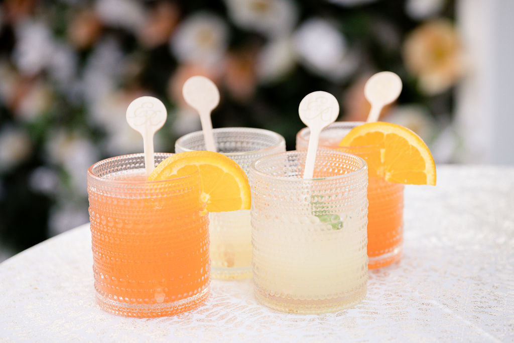 Four textured glasses with two orange and two light yellow drinks sit on a white lace table. Each glass has a round stir stick and a slice of orange garnish. A blurred floral background is visible behind the drinks.