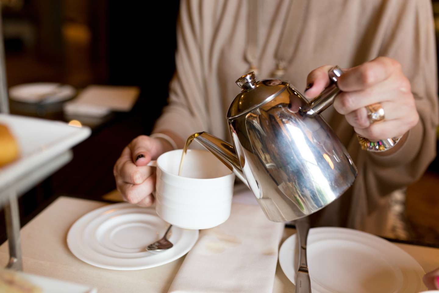 A person wearing a beige top pours tea from a shiny metal teapot into a white cup on a saucer, with a spoon resting inside the cup and plates on the table.