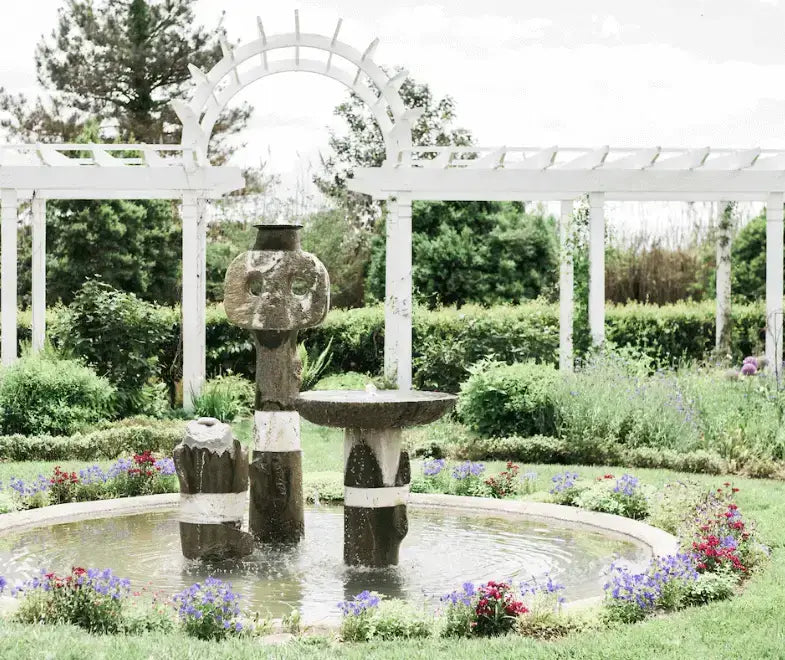 A stone fountain with abstract shapes stands in the center of a circular pond, surrounded by colorful flowers and greenery, with a white pergola and archway in the background.
