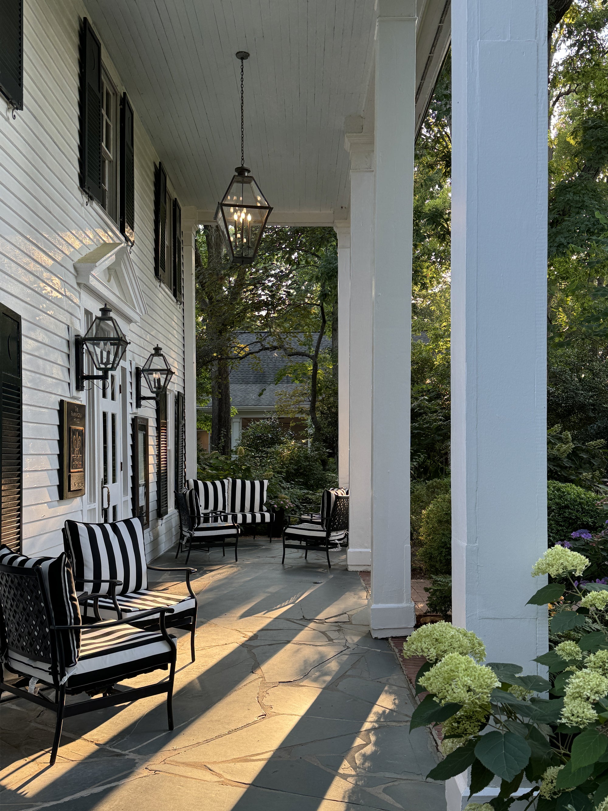 Sunlight streams onto a charming porch with black-and-white striped chairs, hanging lanterns, and white columns, surrounded by lush greenery and blooming hydrangeas, creating a serene, inviting atmosphere.