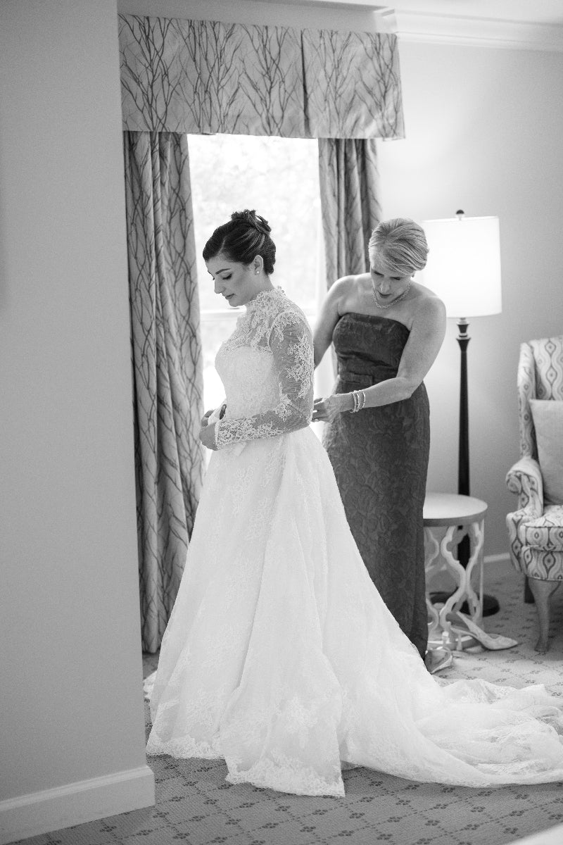 A woman in a lace wedding dress stands as another woman helps adjust the back of her gown in a softly lit room with a window, lamp, and armchair. The scene is captured in black and white.