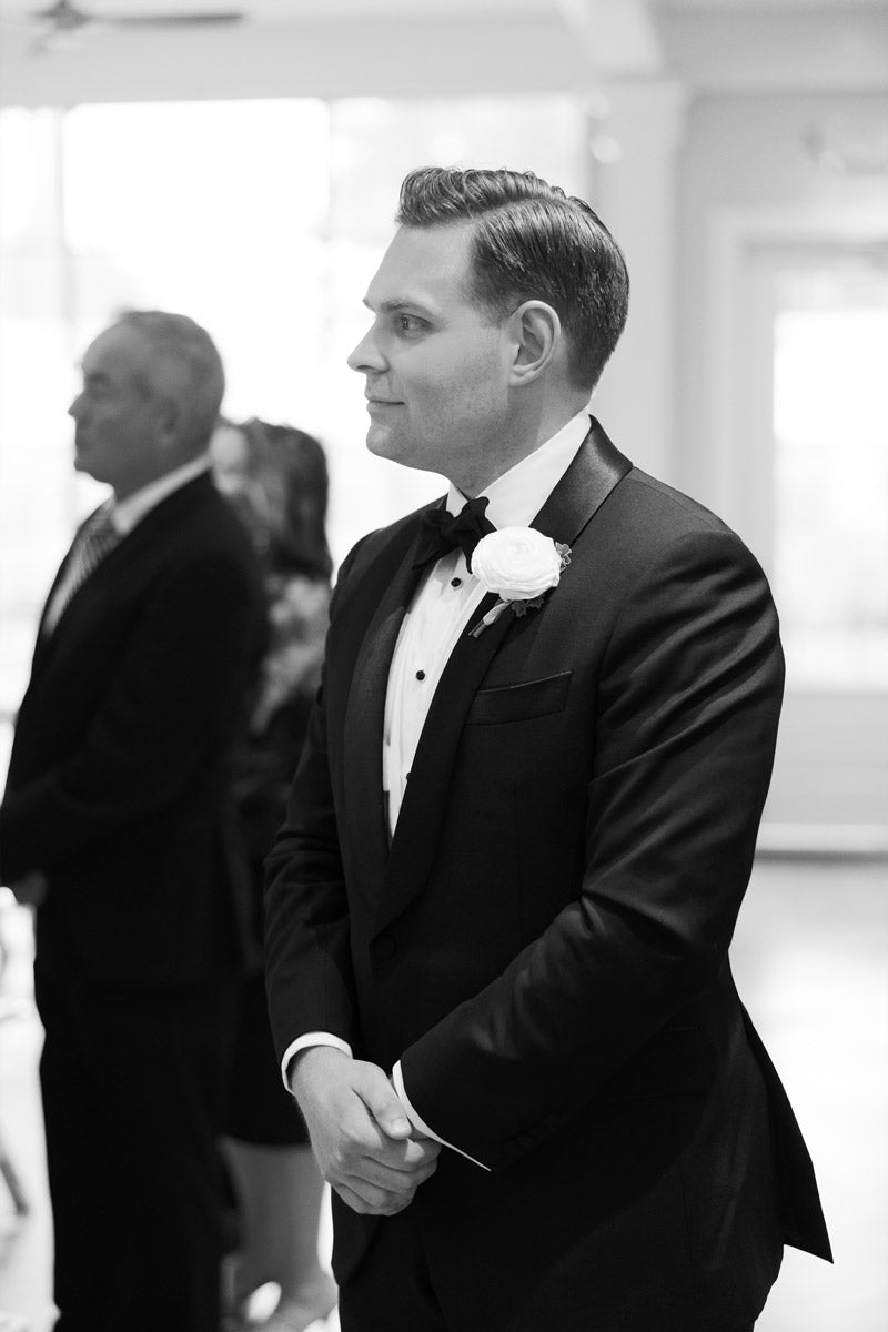 A man in a tuxedo with a white boutonniere stands indoors, looking forward and smiling slightly. Other people, dressed formally, stand behind him. The setting appears to be a wedding or formal event. Black and white photo.