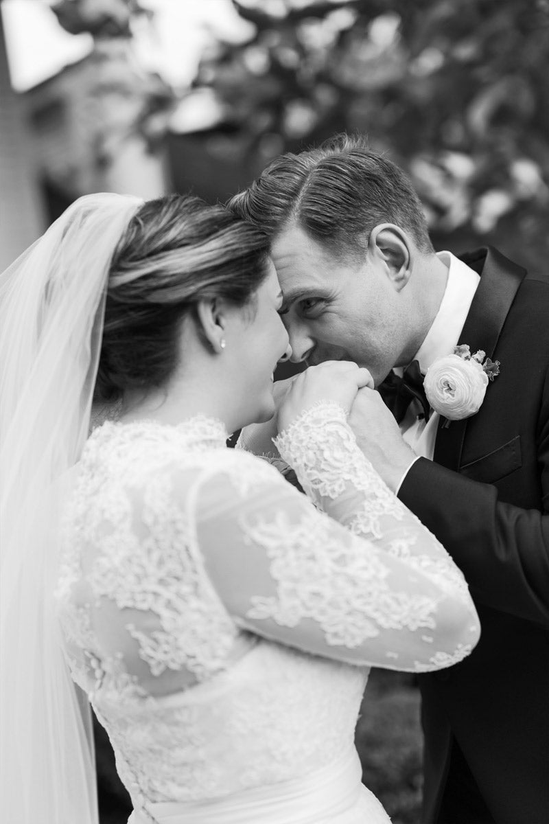 A groom in a suit gently kisses the hand of his bride, who wears a lace wedding dress and veil. They stand close, smiling at each other, sharing an intimate and joyful moment outdoors.