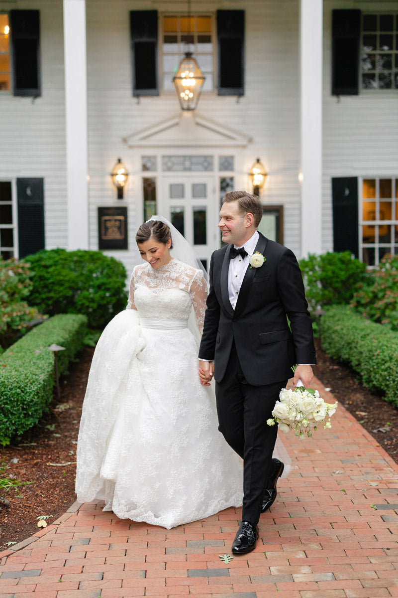 A bride in a white lace wedding dress and a groom in a black tuxedo walk hand-in-hand on a brick path outside a white, colonial-style house. The groom holds a bouquet of white flowers.