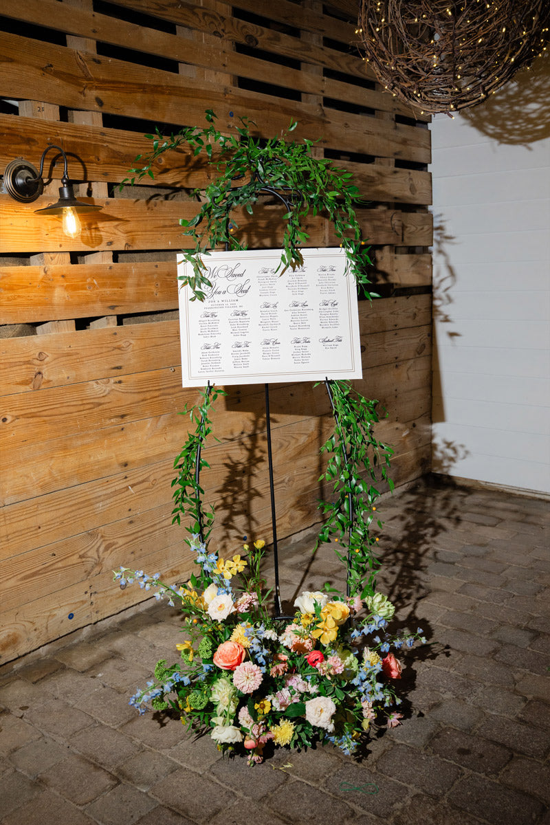 A wedding seating chart displayed on an easel draped with greenery, with a floral arrangement at the base. The setup is against a wooden wall with a hanging light and cobblestone floor.
