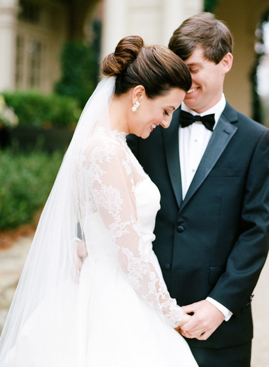 A bride in a white dress with lace sleeves and a long veil stands close to a groom in a black tuxedo. They are smiling, holding hands, and touching foreheads outdoors.