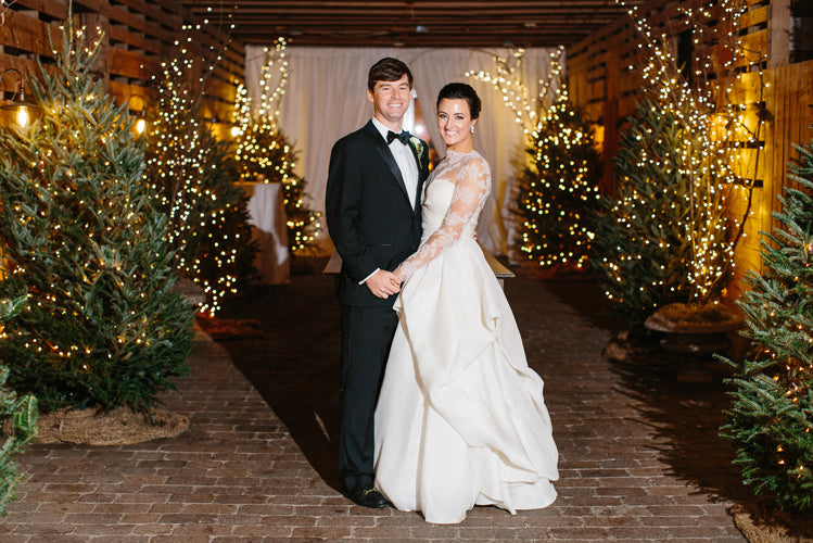 A bride in a white gown and a groom in a black tuxedo smile and pose together in a festive, warmly lit barn decorated with Christmas trees and twinkling string lights.