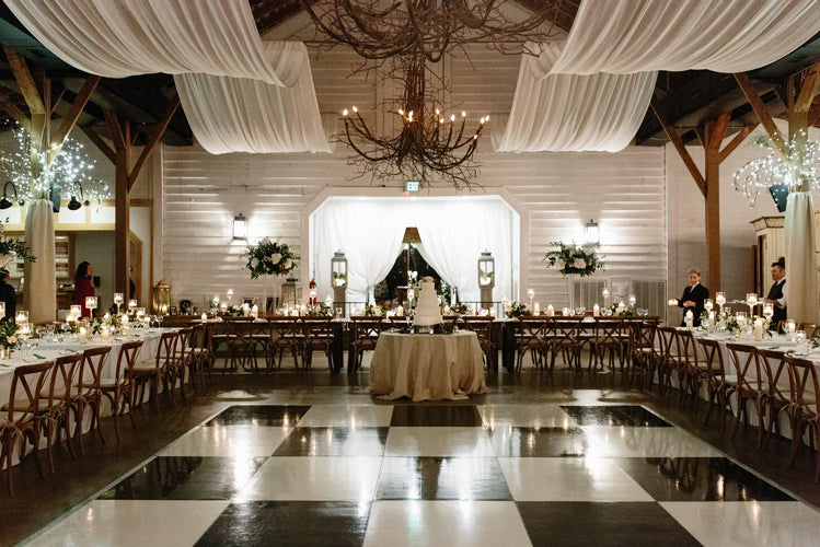 A beautifully decorated wedding reception hall with draped white fabric on the ceiling, a large cake centerpiece on a round table, long dining tables set with candles and flowers, and a black-and-white checkered dance floor.