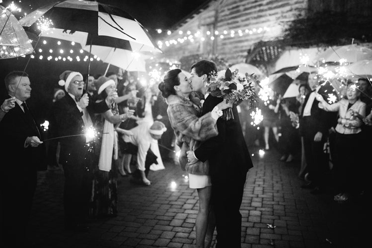 A couple kisses while surrounded by guests holding sparklers and umbrellas at an outdoor nighttime celebration, with string lights and festive attire creating a joyful atmosphere.