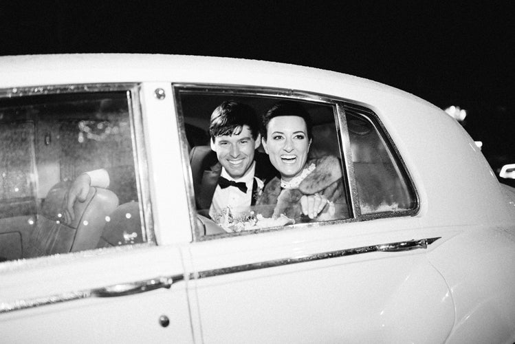 A smiling bride and groom sit together in the backseat of a classic white car, looking out of the open window at night, dressed in formal wedding attire.