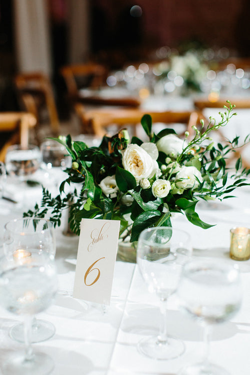 A close-up of a wedding table centerpiece with white flowers and greenery, surrounded by glassware and candles. A table card with “Table 6” in elegant gold script is placed in front of the arrangement.