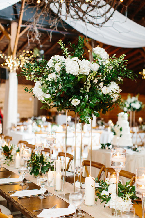 Elegant wedding reception table with a tall floral centerpiece of white flowers and greenery, surrounded by candles, glassware, and decorated wooden chairs in a warmly lit rustic venue with hanging branches and string lights.