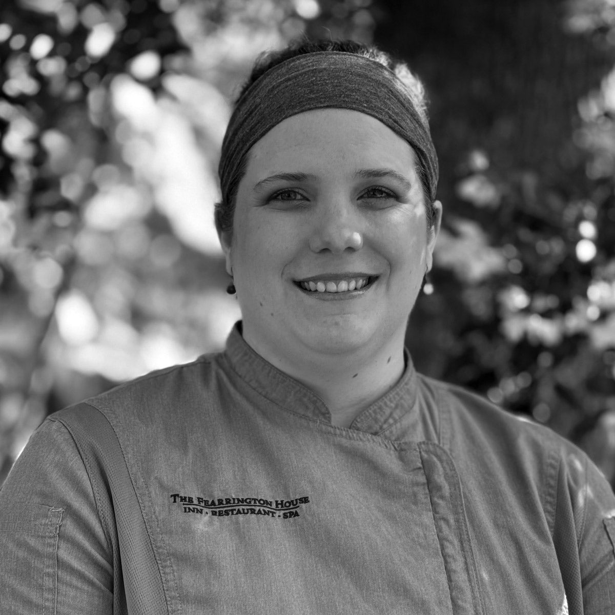 A person wearing a headband and a chef’s jacket stands outdoors, smiling. The jacket is embroidered with “The Peabody House Inn Restaurant, Spa.” Trees and sunlight are blurred in the background. The image is in black and white.
