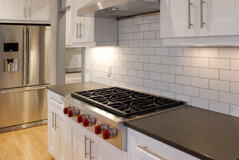 Modern kitchen with stainless steel appliances, a large refrigerator, white cabinets, subway tile backsplash, black countertops, and a gas stove with red knobs under a range hood. Bright, clean, and well-lit space.