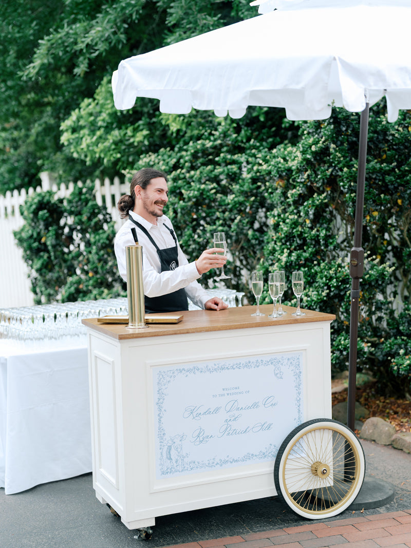 A smiling server in a white shirt and black apron stands behind a mobile drink cart with glasses of champagne, under a white umbrella, outdoors near greenery and a white picket fence.