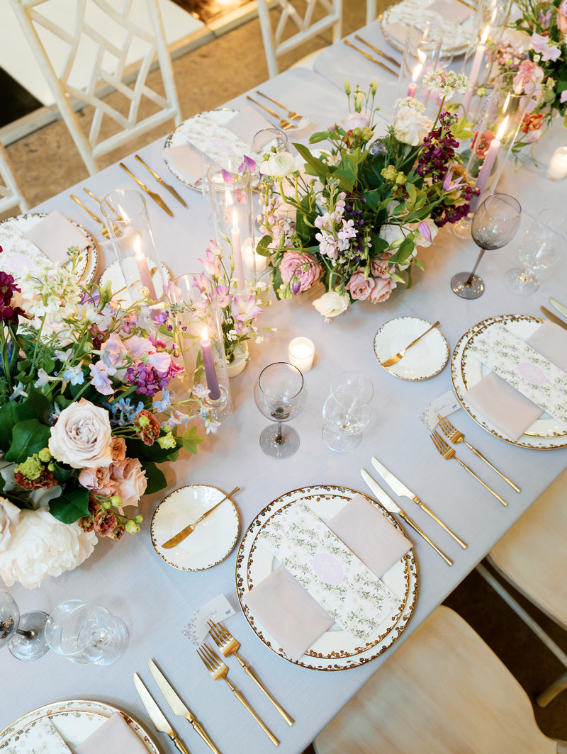 An elegant table setting with floral-patterned plates, gold cutlery, pink napkins, tall purple candles, and lush floral centerpieces featuring pink, purple, and white flowers on a light tablecloth.