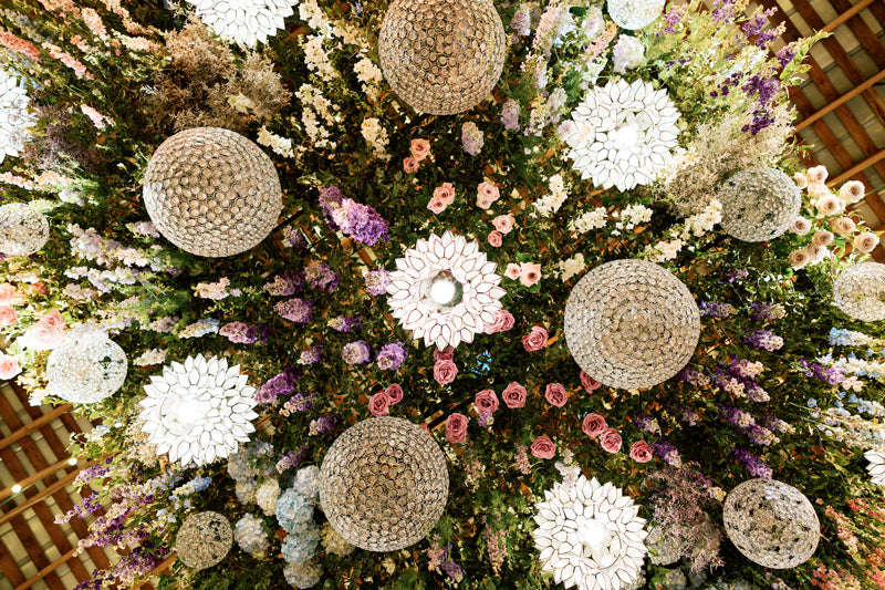 A decorative ceiling arrangement featuring large spherical lights and white floral patterns surrounded by an abundance of colorful flowers, including roses and greenery.