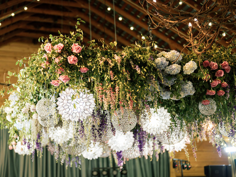 A lush, floral chandelier hangs from a wooden ceiling, adorned with pink roses, white hydrangeas, trailing greenery, and decorative white lanterns, creating an elegant, garden-like atmosphere indoors.