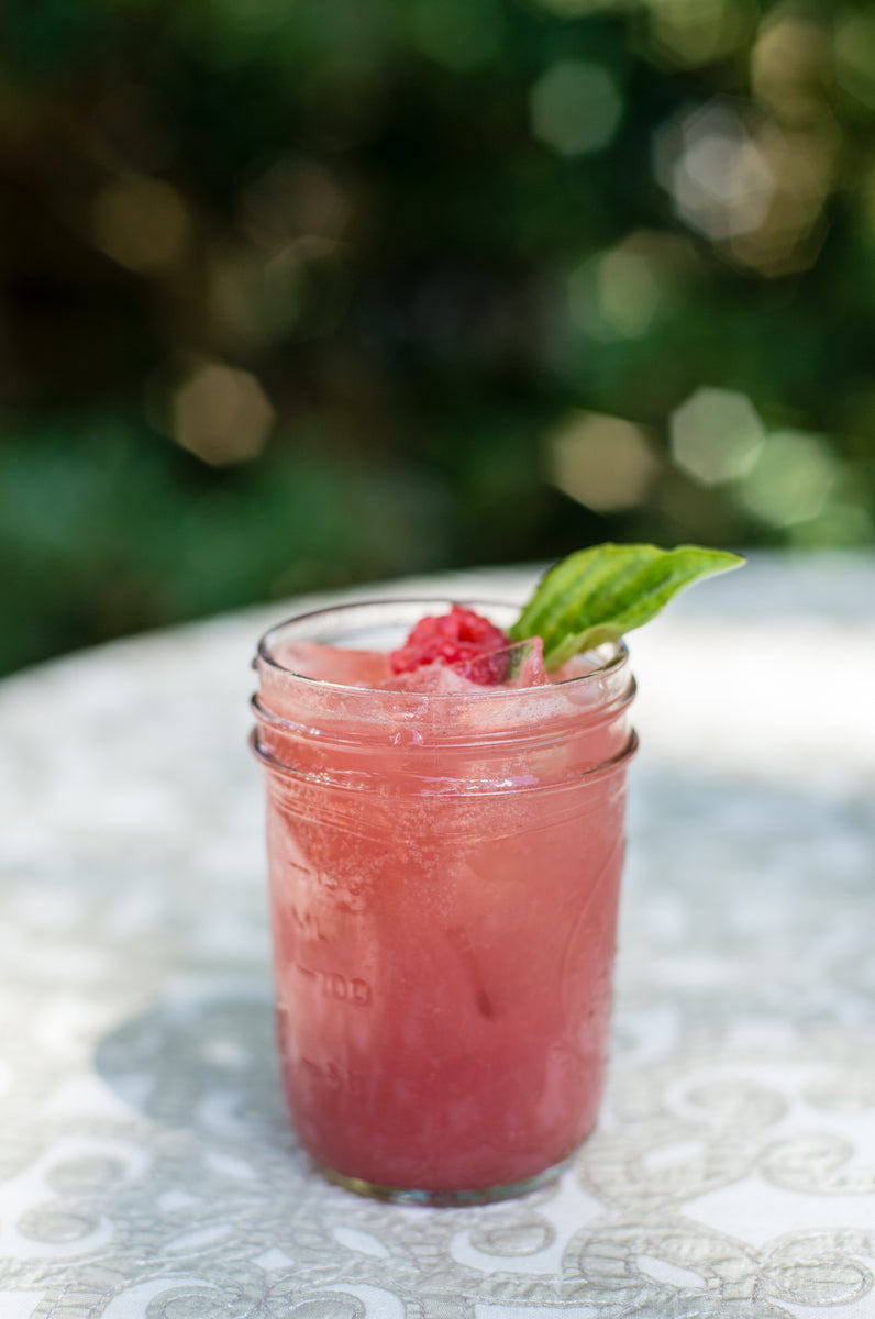A pink iced drink in a mason jar, garnished with a raspberry and a fresh green leaf, sits on a patterned white table with a blurred green background.