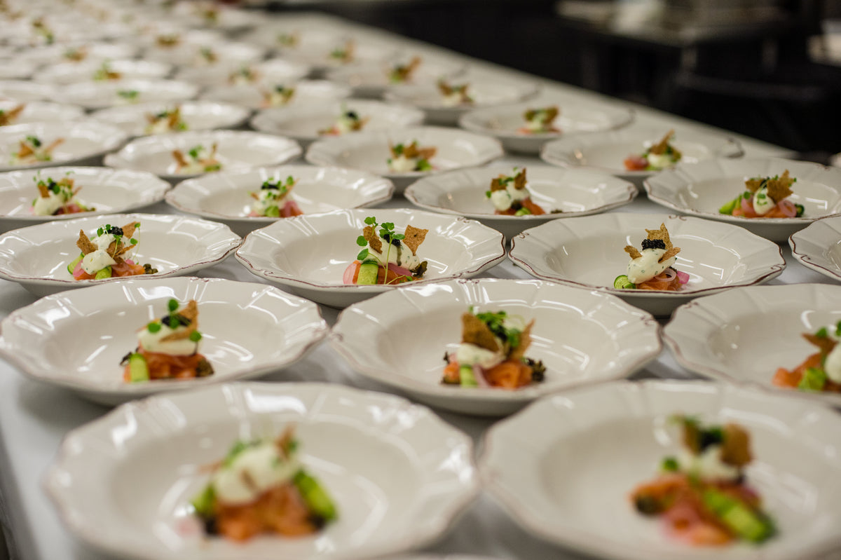 Rows of white plates with gourmet appetizers, garnished with herbs and arranged neatly on a long table, ready for a large event or banquet.