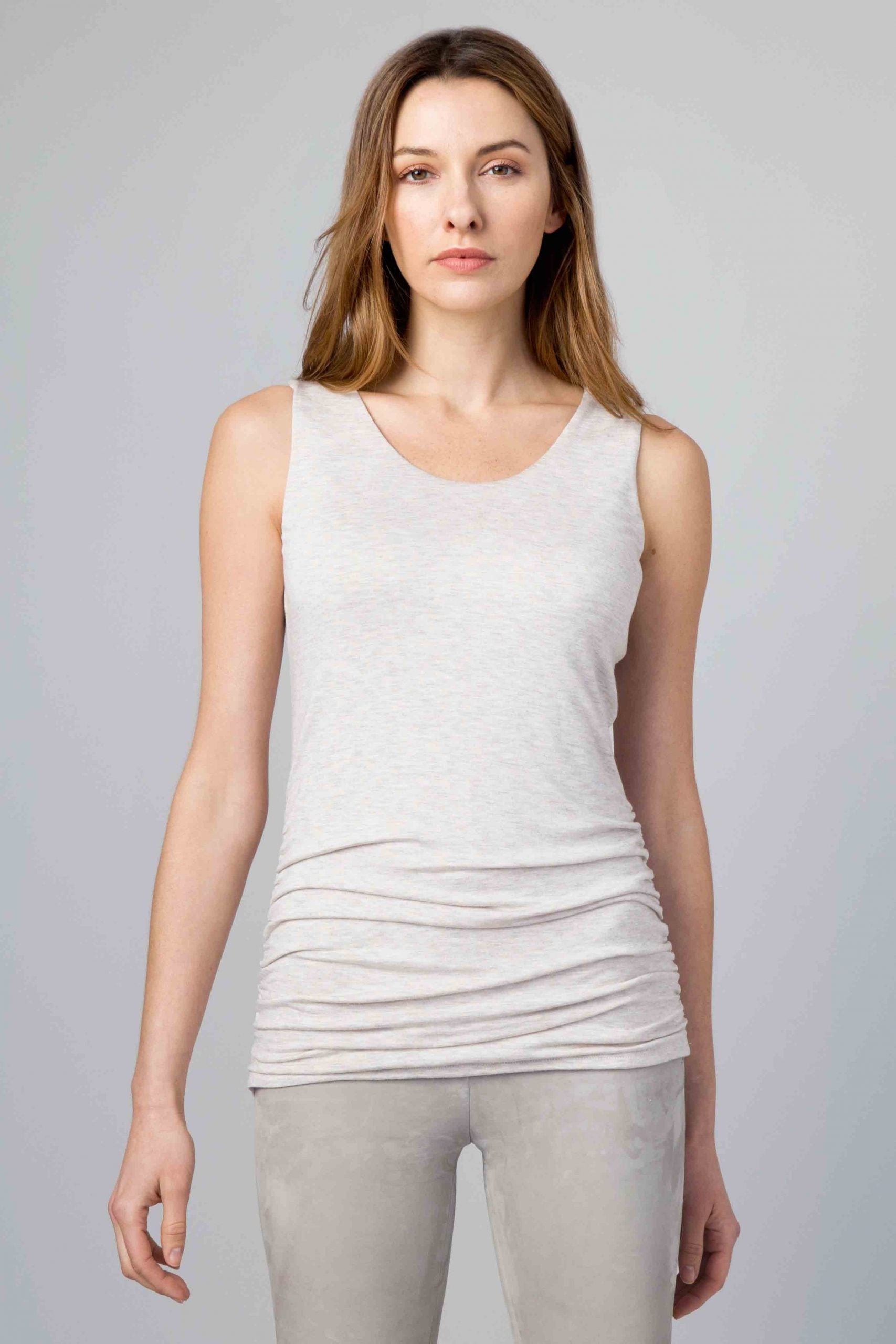 A person with long light brown hair is standing against a plain gray background. They are wearing a light gray sleeveless top, specifically the KINROSS CASHMERE - RUCHED COTTON TANK IN DUSK by KINROSS, and light gray pants which are likely part of Kinross Clothing's elegant collection. The person's expression is neutral, and they have a relaxed posture with their arms by their sides.