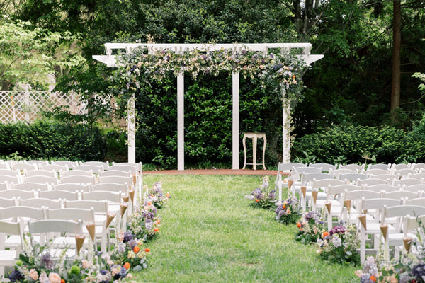 An outdoor wedding ceremony setup with rows of white chairs facing a white pergola adorned with flowers. The aisle is lined with floral arrangements, and lush greenery surrounds the area.
