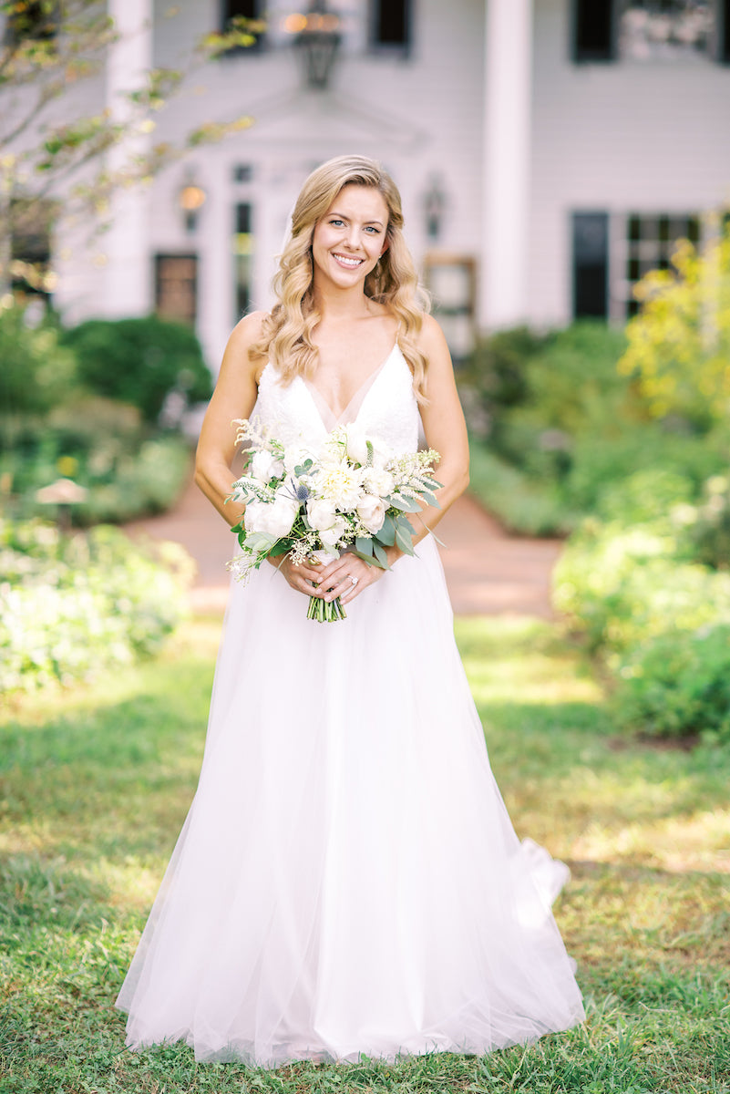 A smiling bride in a white dress holds a bouquet of white flowers, standing outside on a grassy path with a white house and greenery in the background.